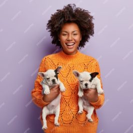 plan-femme-joyeuse-peau-sombre-aux-cheveux-boucles-tient-deux-nouveaux-chiots-mignons-race-trouve-nouvel-hote-pour-animaux-compagnie-est-bonne-humeur-porte-pull-orange-isole-mur-violet_273609-26251